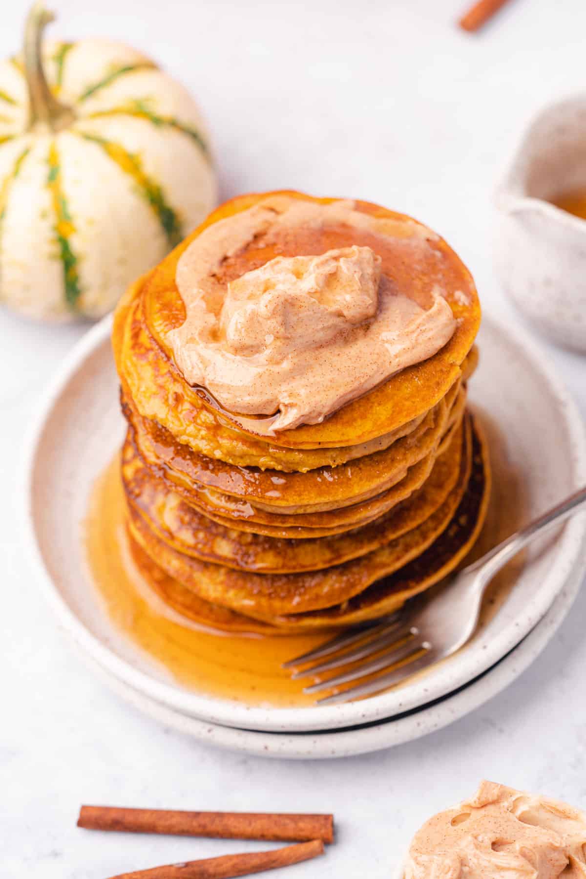 stack of bright orange pumpkin pancakes topped with spiced butter, with cinnamon sticks and gourds surrounding the plate and syrup dripping down the stack. 