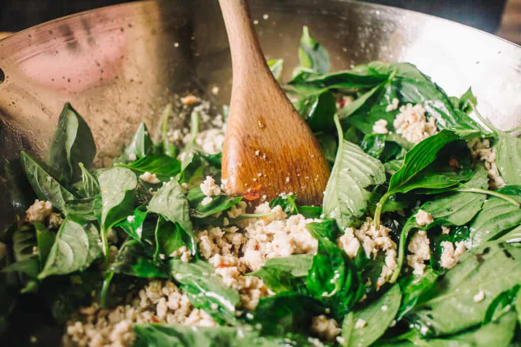 wooden spoon stirring in fresh thai basil leaves in a wok with cooked chicken