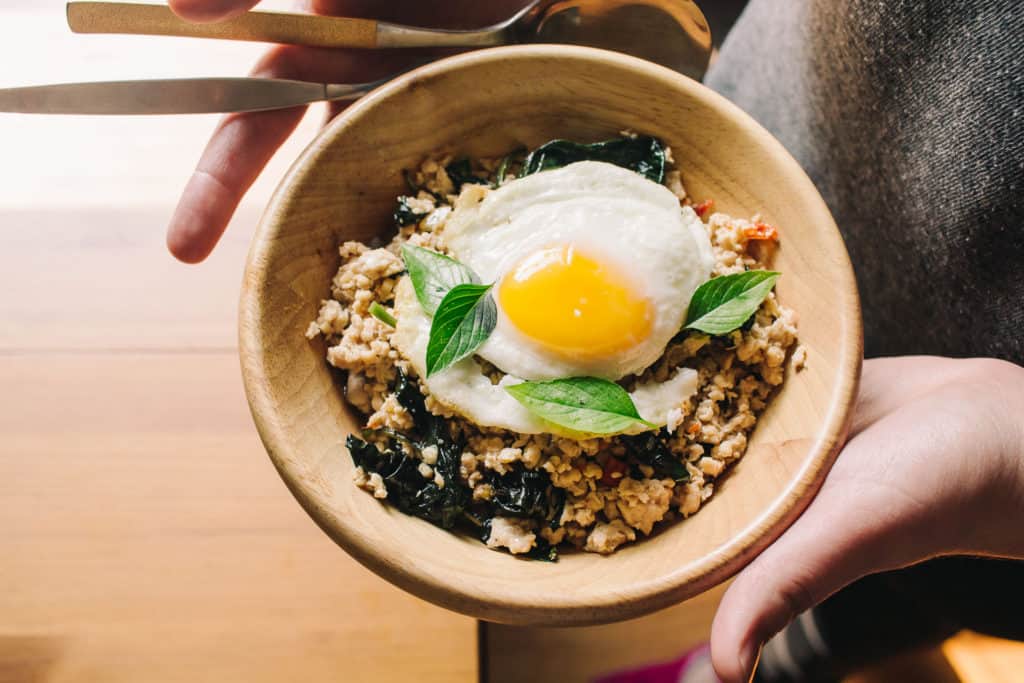 hands holding a bamboo bowl with thai basil chicken in it with an egg on top