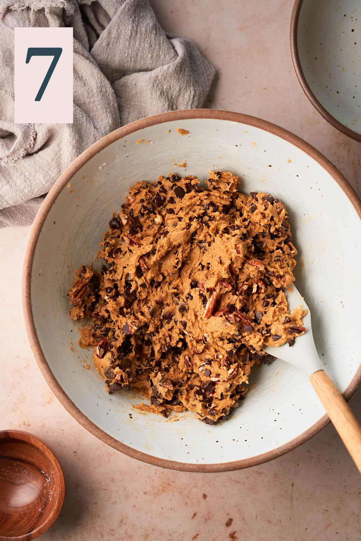 cookie dough with chocolate chips and pecan pieces in a large mixing bowl with a rubber spatula folding it all together. 