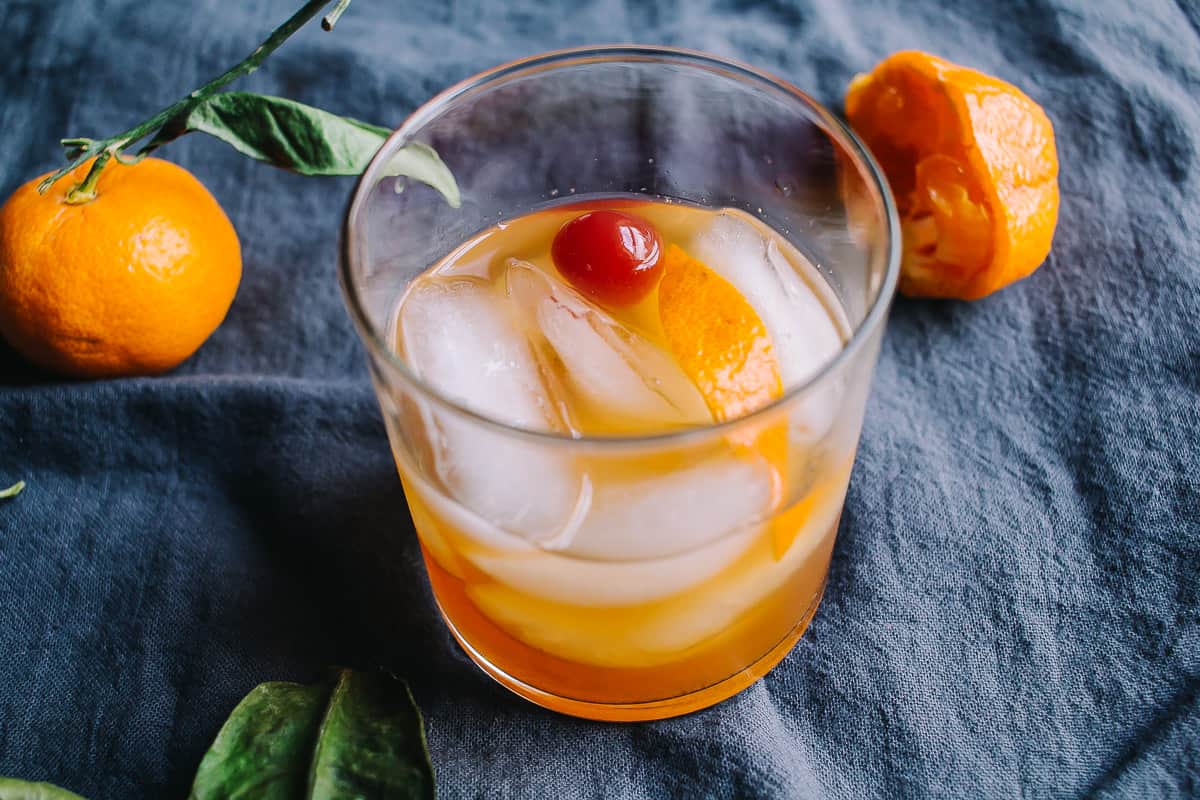 tangerine old fashioned on a grey napkin with tangerines with stems and leaves surrounding it