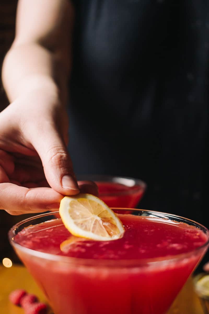 hand coming out from a black background placing a lemon slice on top of a raspberry and pomegranate cocktail