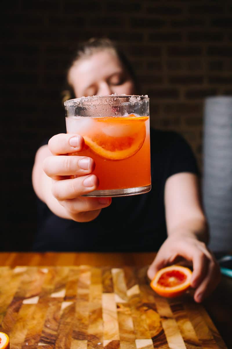 girl holding a blood orange cocktail in one hand toward the camera and a half of a blood orange in another hand