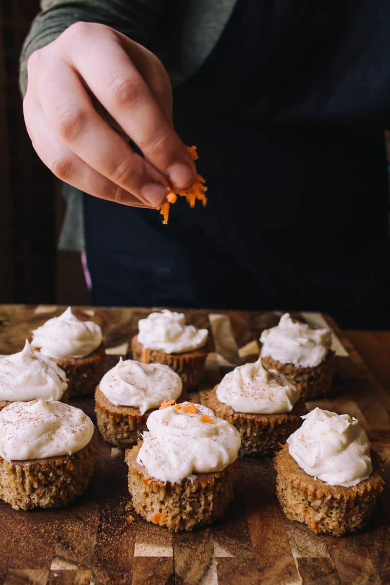 hand dropping shreds of grated carrots on top of carrot cake cupcakes