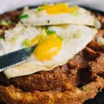 Two crispy savory waffles topped with eggs and green onions with a knife cutting into the yolk