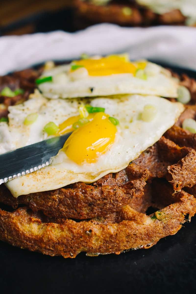 Two crispy savory waffles topped with eggs and green onions with a knife cutting into the yolk