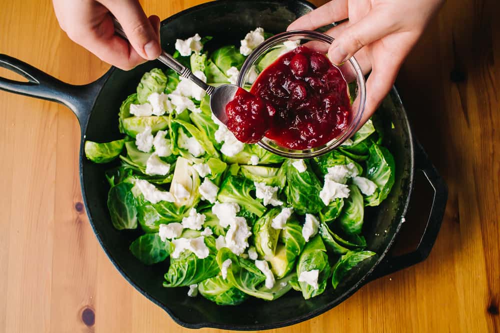 hand with bowl spooning cranberry sauce into Brussel sprout skillet