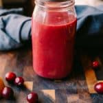 jar of Sugar Free Cranberry Lemon Vinaigrette in a jar on a brown chopping block with cranberries and a grey napkin surrounding it