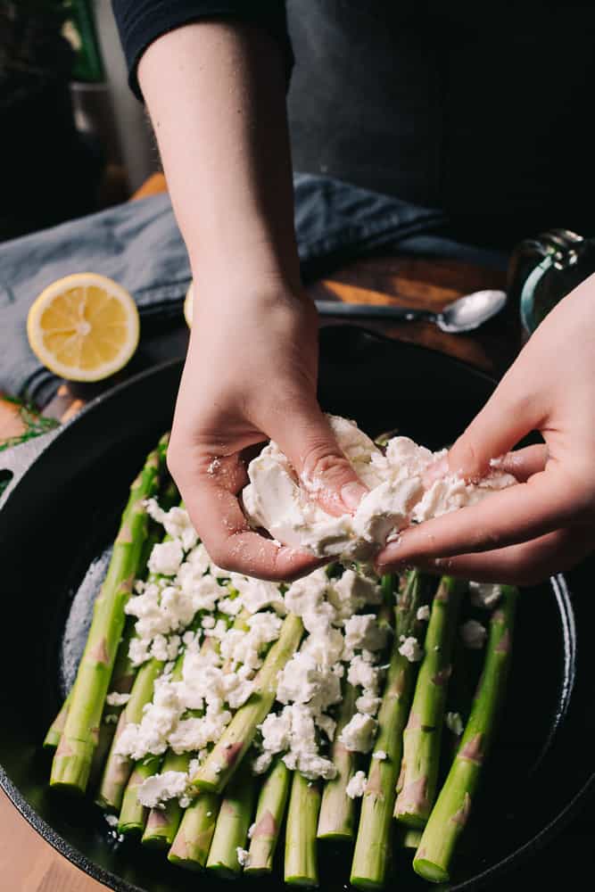hands crumbling goat cheese on a cast iron with asparagus