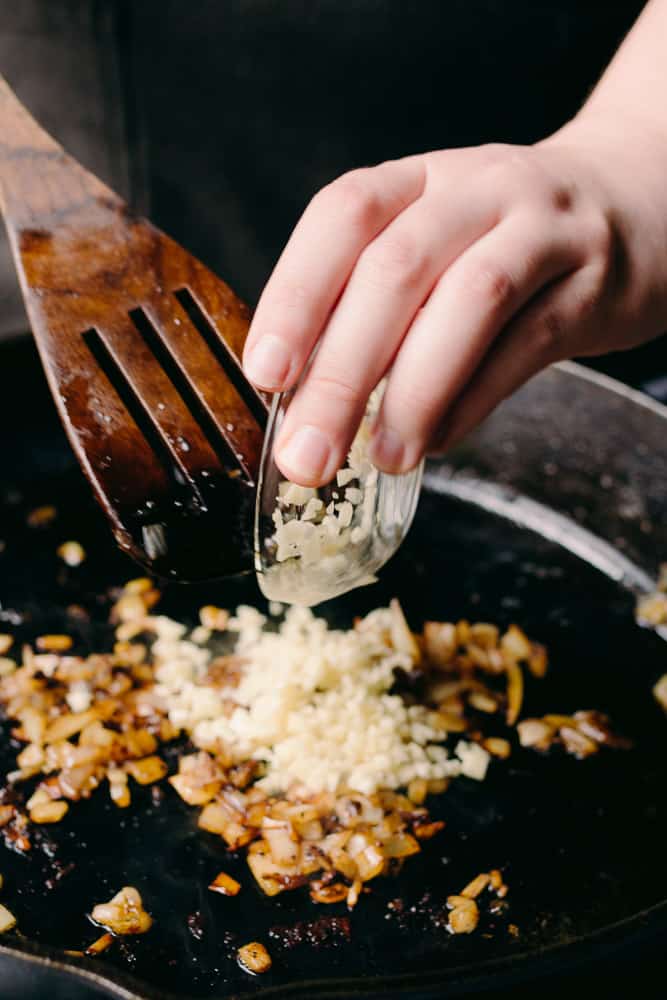 hand adding garlic to cooked onions in a cast iron skillet
