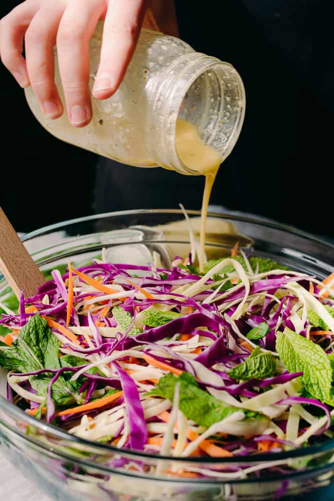 pouring honey lemon vinaigrette dressing on top of purple cabbage salad with carrots and mint