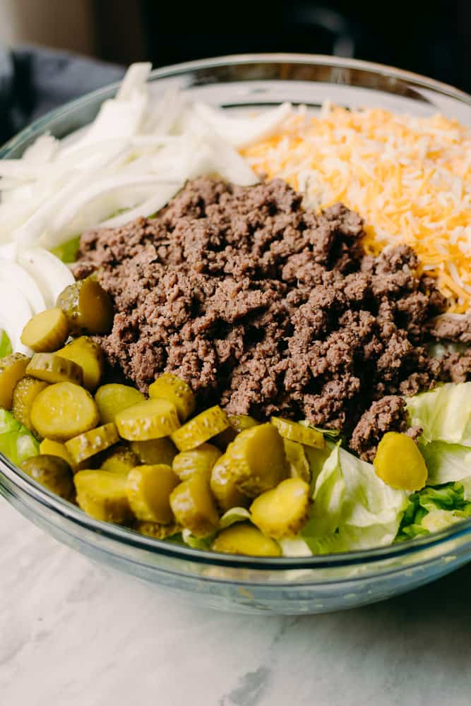 lettuce ground beef pickles onions shredded cheese in a glass bowl of healthier cheeseburger salad