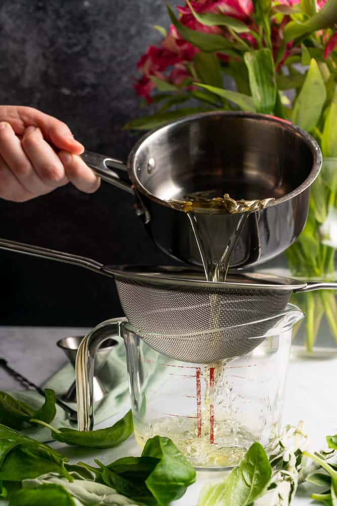 hand pouring herbed sugar free simple syrup from a small sauce pan through a strainer into a measuring glass
