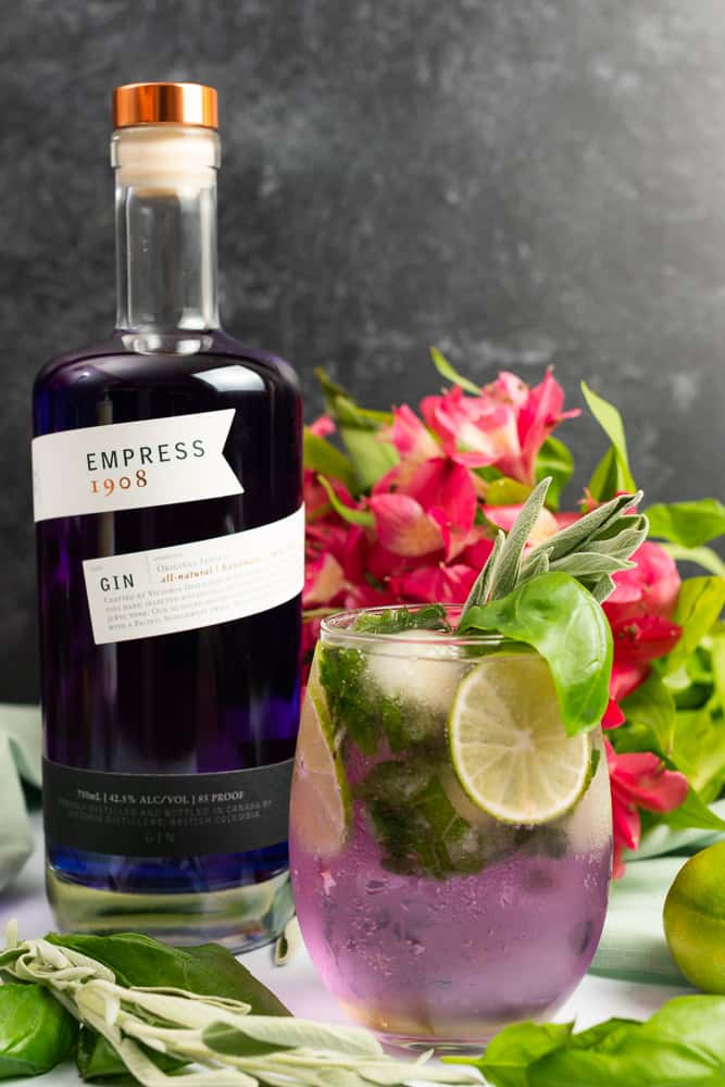 summer herb gin and tonic made with empress gin with a bottle beside the cocktail and pink flowers in the background