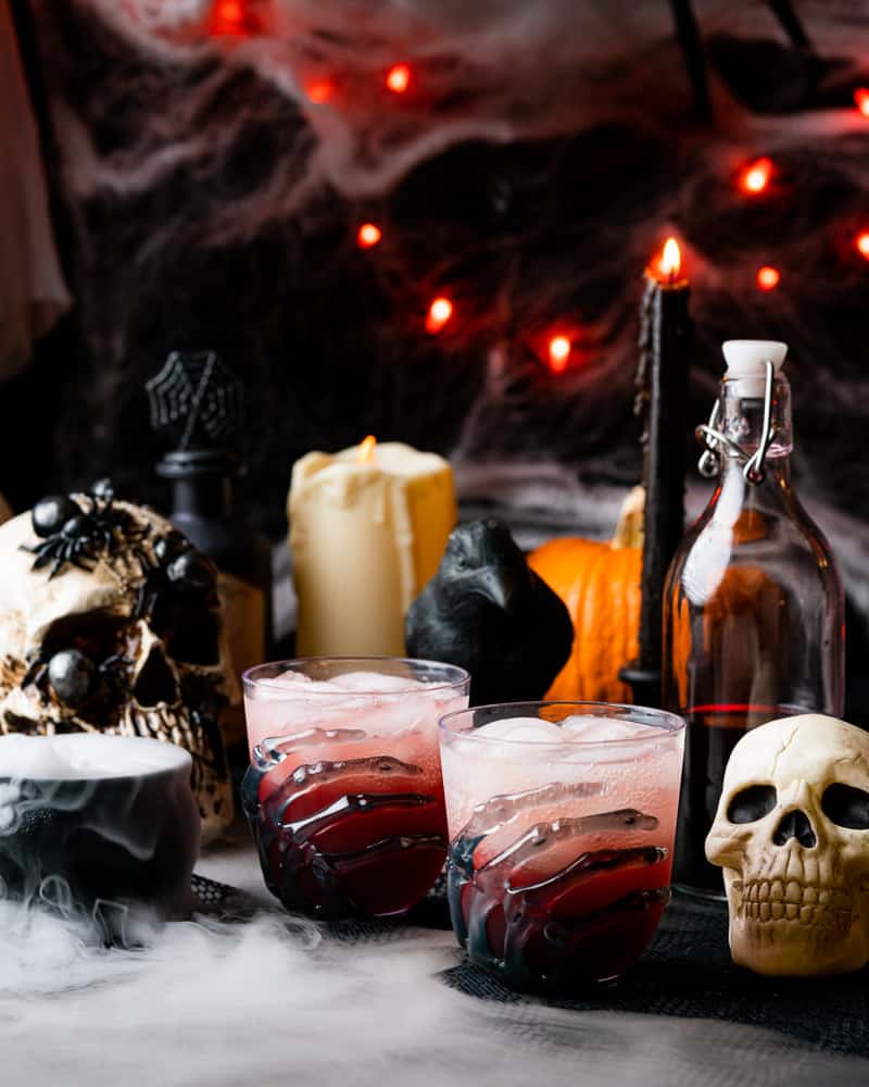 a halloween scene with two red cocktails with skeleton hands skulls pumpkins candles a raven and dry ice with orange lights in the background