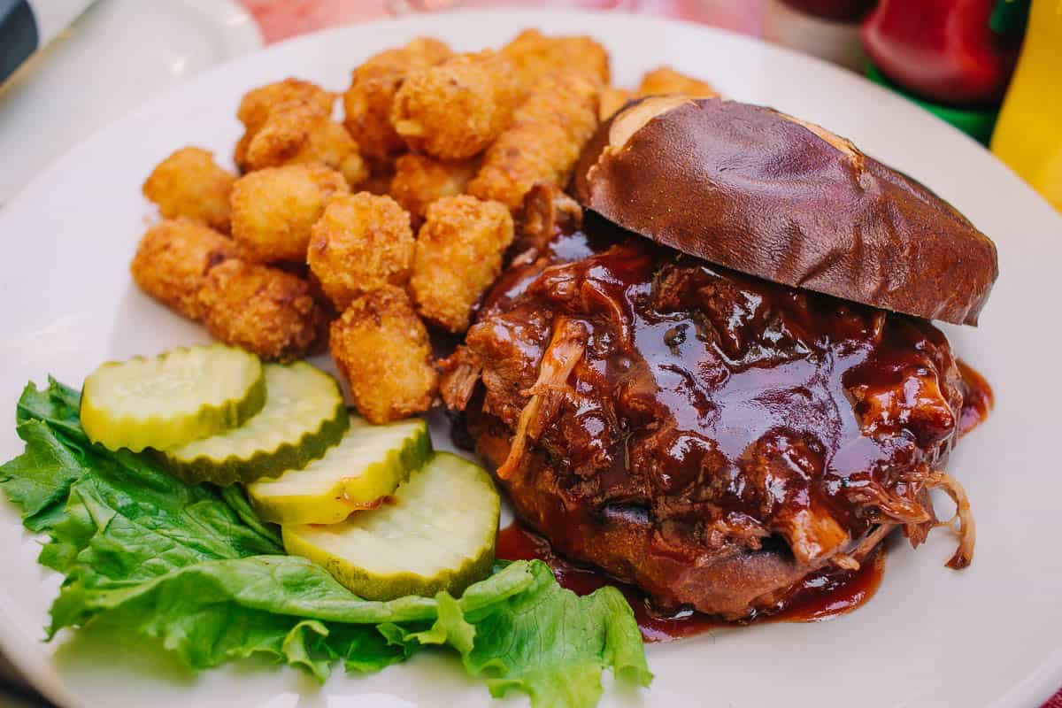 smothered pork bbq sandwich on a pretzel bun with tater tots and pickles
