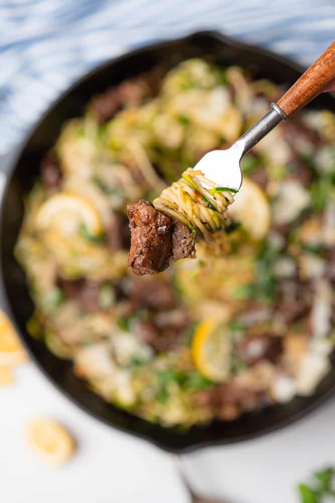 forkful being up held up by a hand of zucchini noodles with a bite of juicy steak on the end