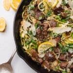 garlic butter steak bites with lemon zucchini noodles with a big metal serving spoon and lemons in a cast iron skillet