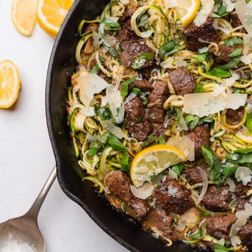 garlic butter steak bites with lemon zucchini noodles with a big metal serving spoon and lemons in a cast iron skillet