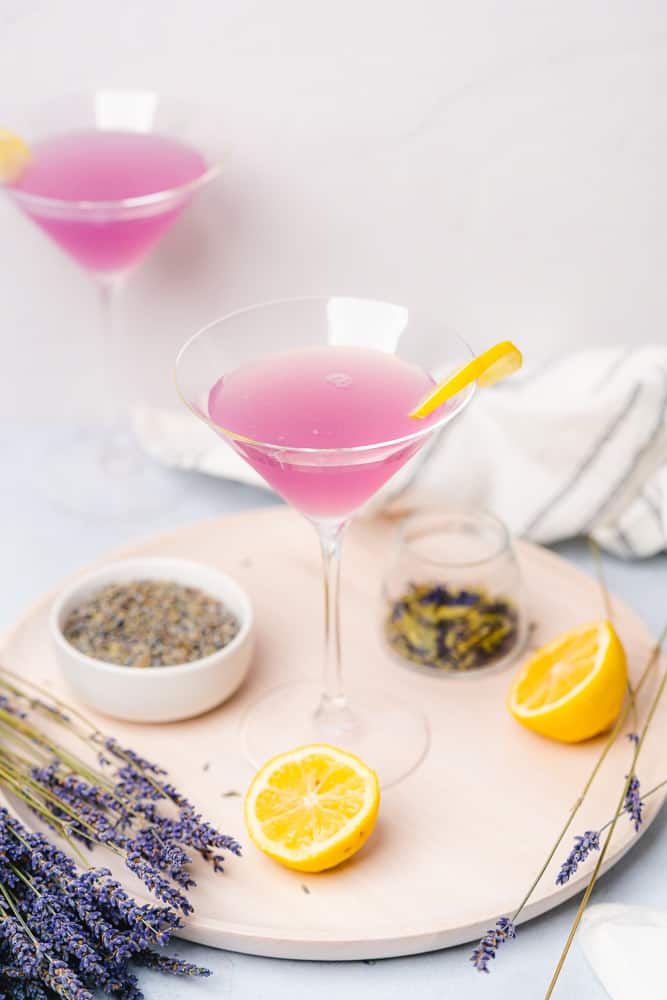 lavender lemon martini on a white wooden board with whole lemons and dried butterfly pea flowers and lavender buds