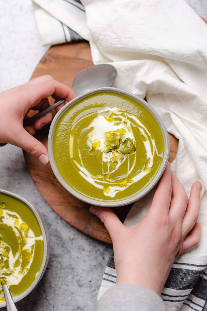 two hands grabbing a bowl of potato leek kale soup drizzled with cream and topped with leeks