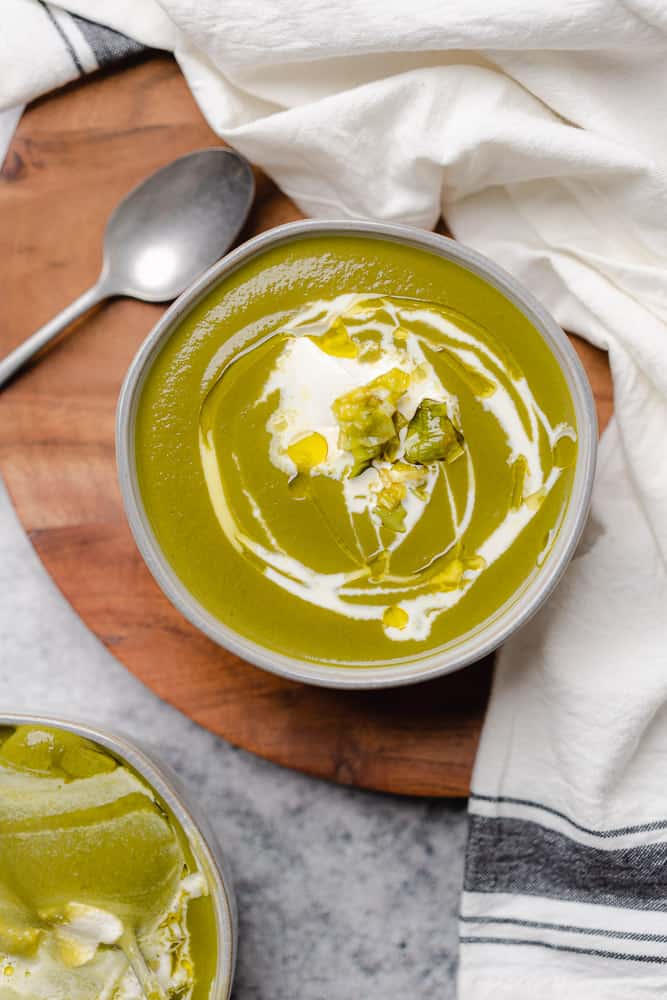 bowl of bright green soup on a wooden background with a drizzle of heavy cream olive oil and sour cream
