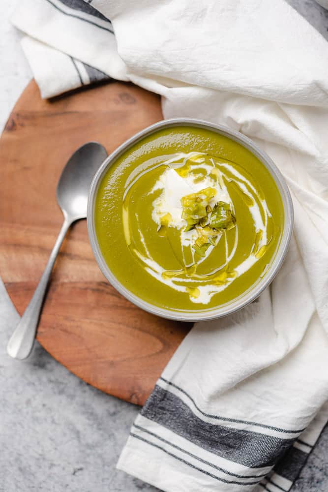 bowl of green soup with drizzle of cream and a white napkin on a wooden back drop