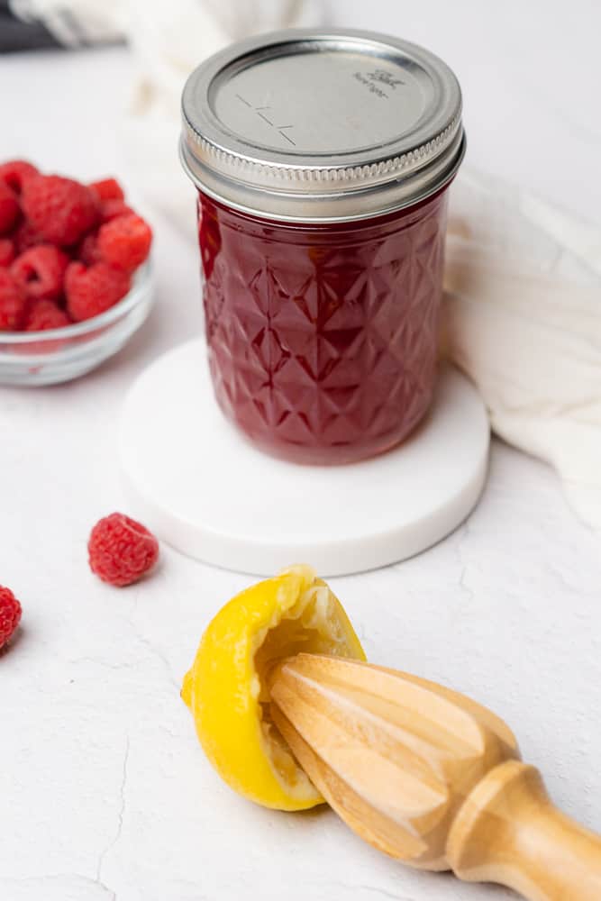 raspberry syrup in a jar surrounded by fresh raspberries, and a lemon that has been reemed