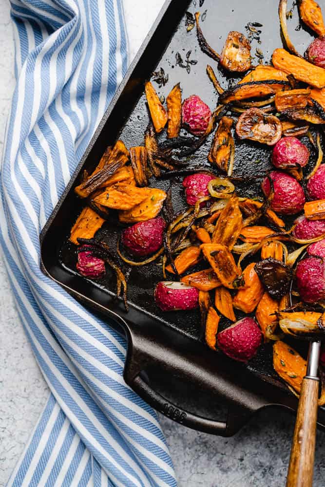 roasted radishes and carrots with onions on a cast iron sheet pan with a blue and white striped napkin 