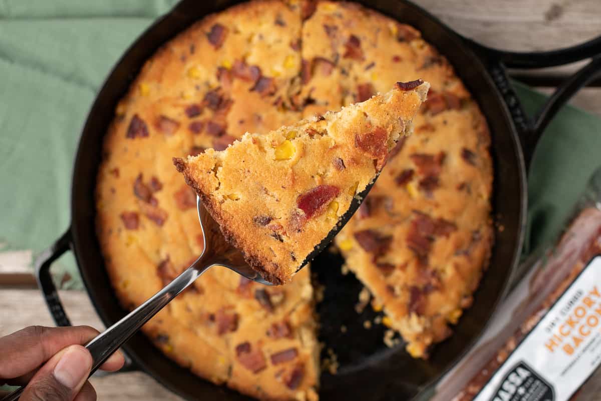 brown butter cornbread with bacon in a cast iron skillet with a slice taken out on a pie serving utensil