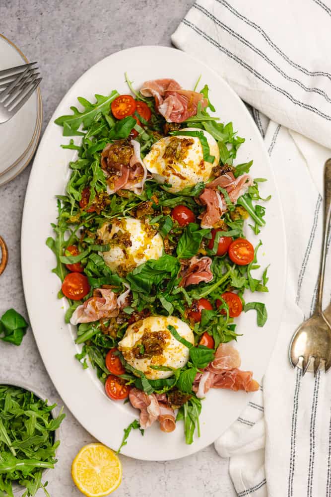 burrata and prosciutto salad with arugula and tomatoes on a white oval platter