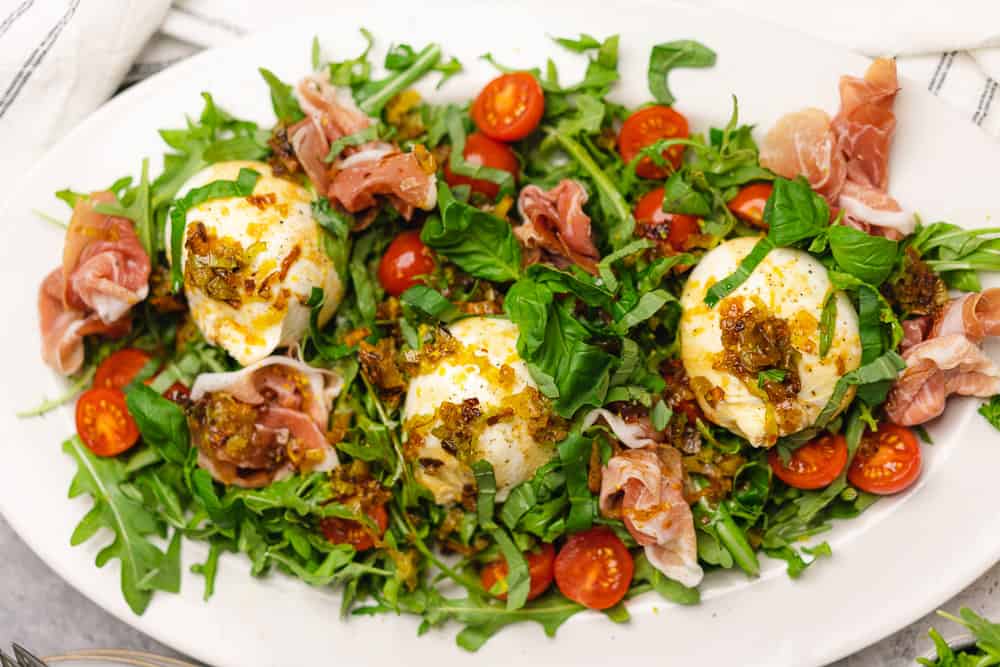 salad with burrata cheese tomatoes balsamic fried leeks lemon and prosciutto on a white oval platter