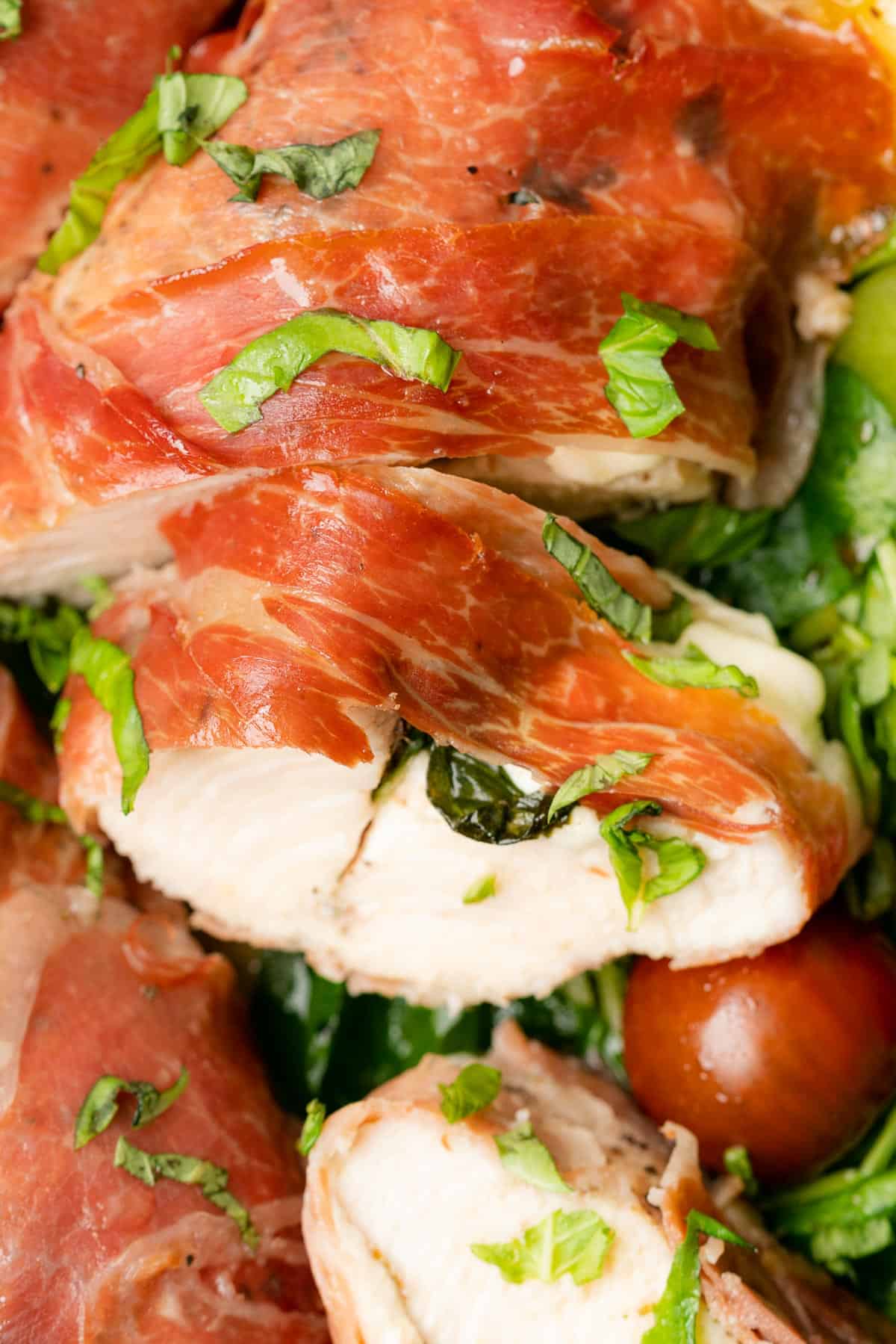 very close up shot of chicken breast topped with prosciutto surrounded by arugula and tomatoes