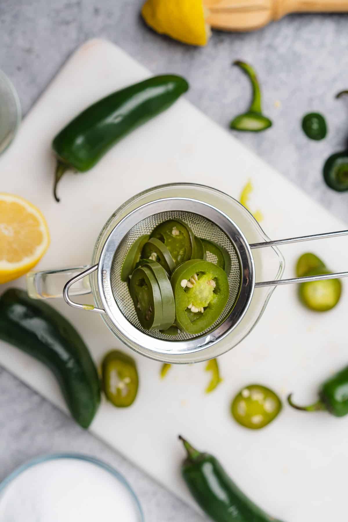 straining away jalapenos from water using a metal sieve with fresh jalapenos and lemon surrounding it 
