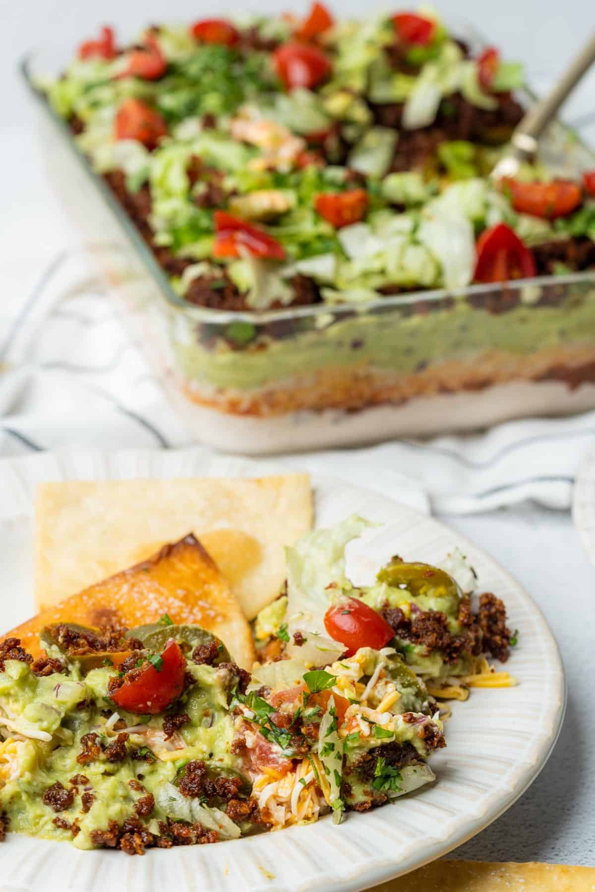 keto 7 layer dip in a glass casserole dish with a plate of dip and low carb tortilla chips in the foreground