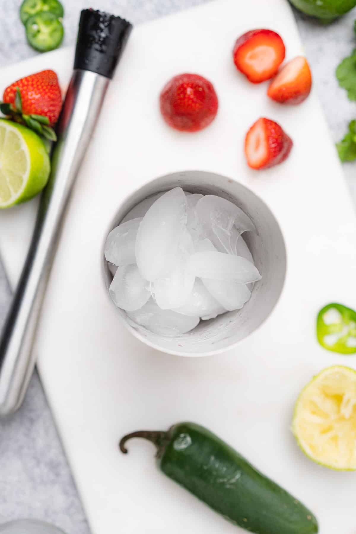ice in a cocktail shaker surrounded by fresh limes strawberries and jalapenos