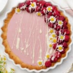stunning light pink raspberry tart topped with fresh raspberries chamomile flowers and a white chocolate drizzle