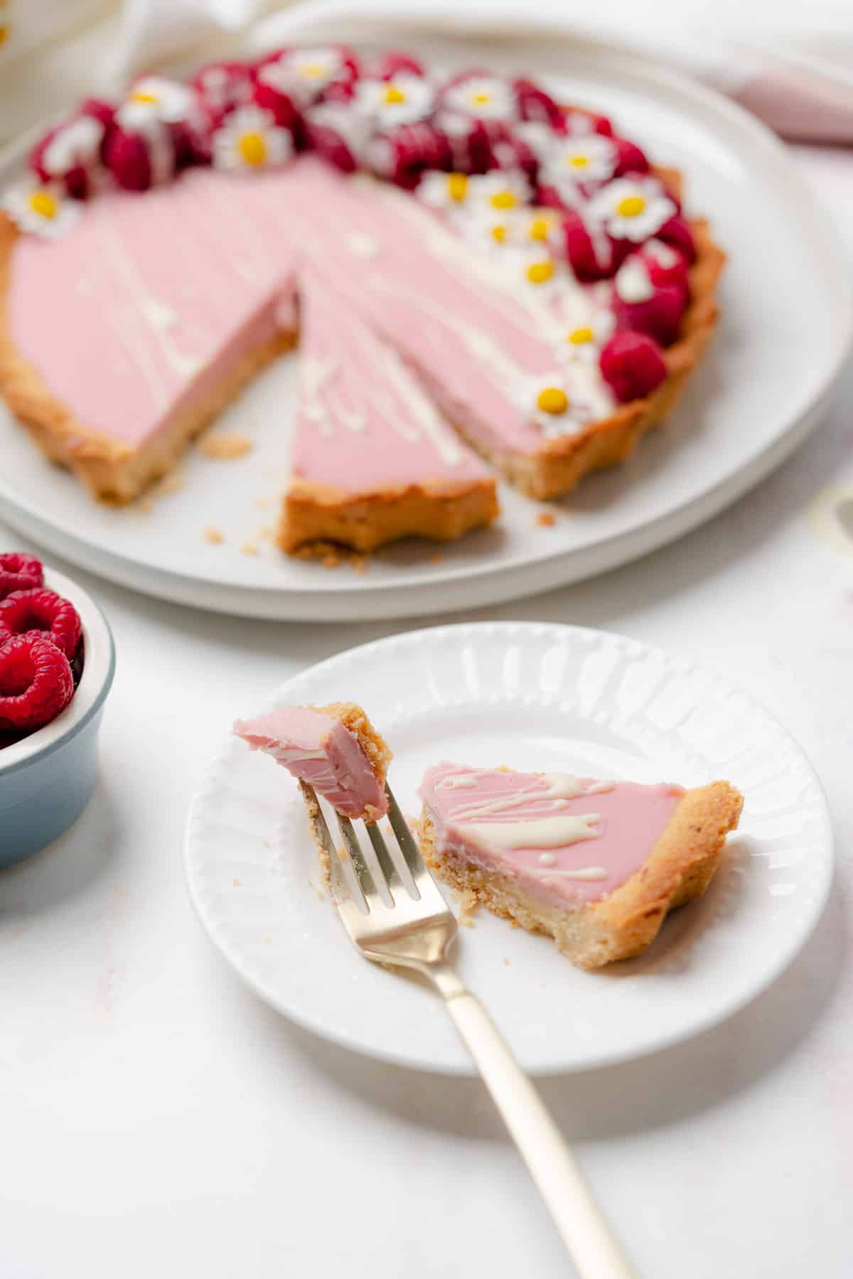 white chocolate raspberry tart on a plate with a slice taken out with a gold fork on another plate