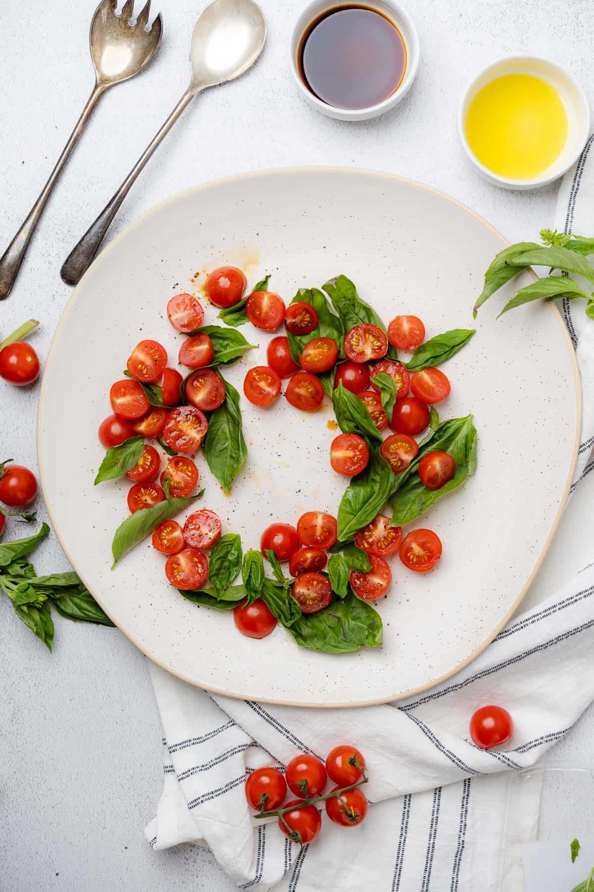 basil and tomatoes in a ring on a white ceramic serving platter surrounded by olive oil and balsamic with a white and blue striped linen napkin
