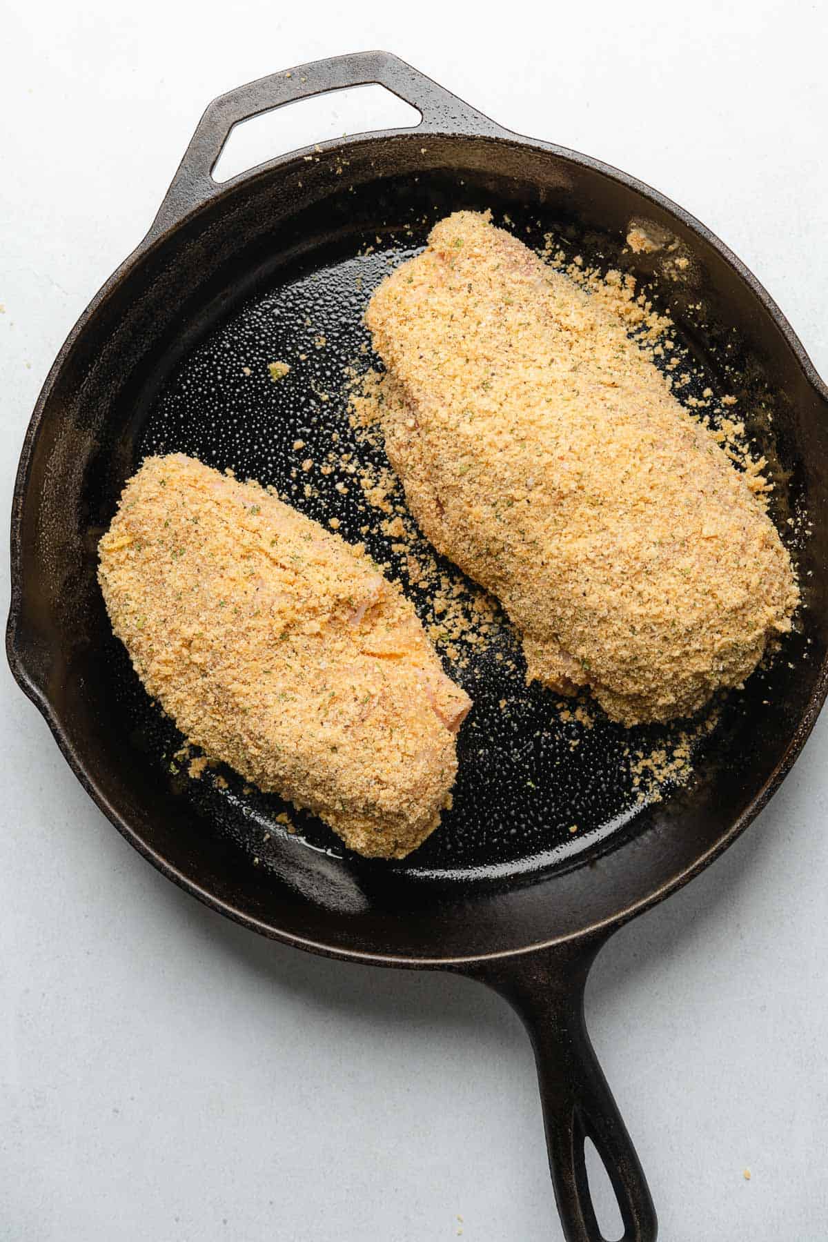 raw chicken breasts coated in pork rind crumbs in a cast iron skillet