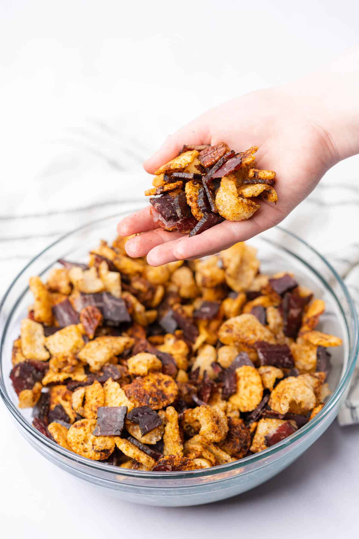 hand held shot of keto snack mix with biltong over a large bowl of party mix