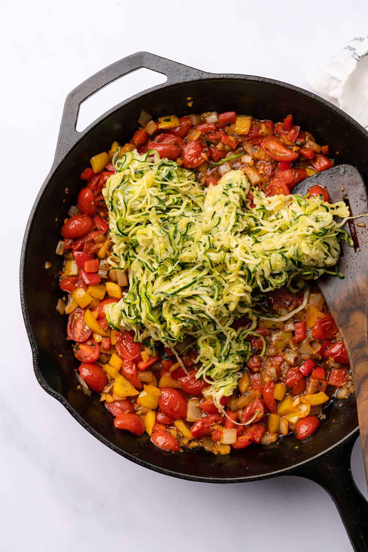 adding zucchini noodles to a skillet with peppers, tomatoes and onions