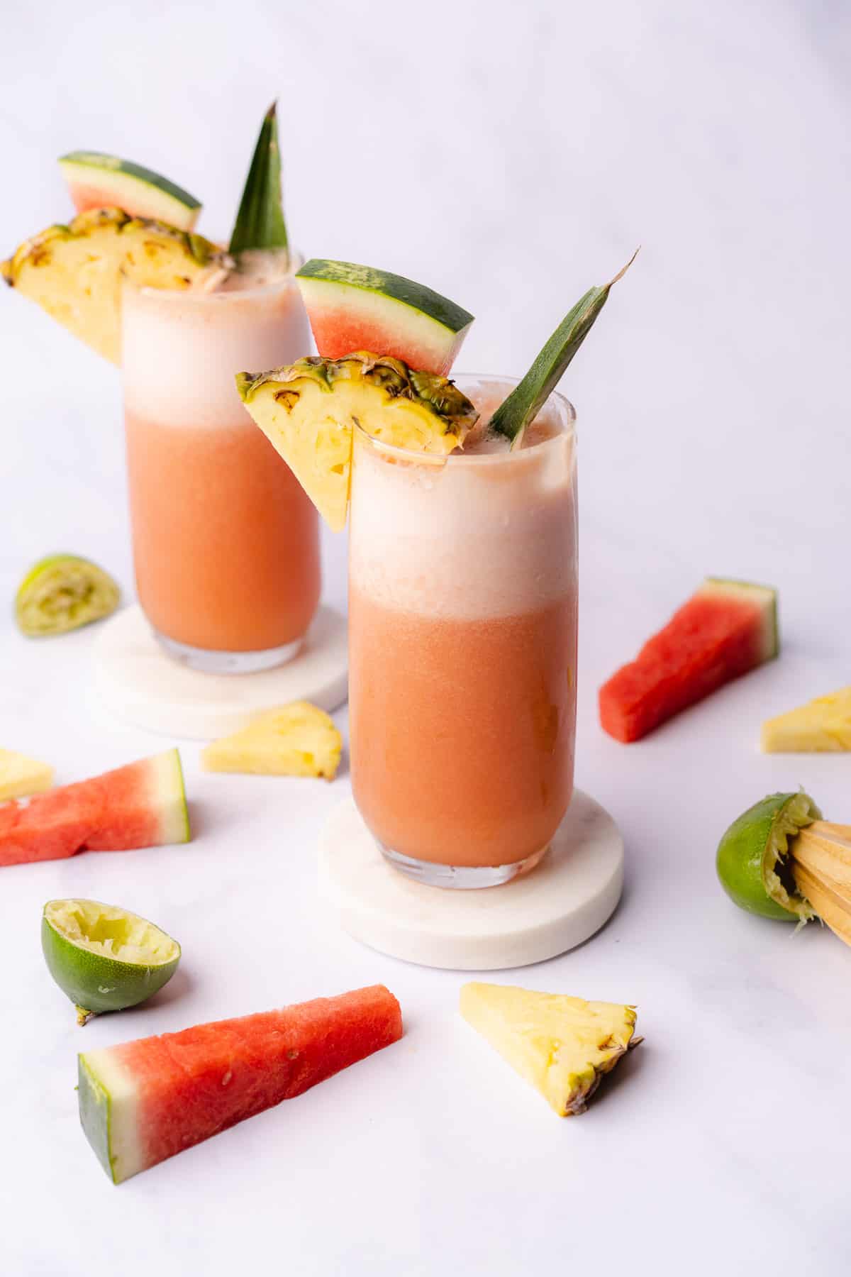two glasses of pineapple watermelon daiquiri with lime and fruit wedges around it 