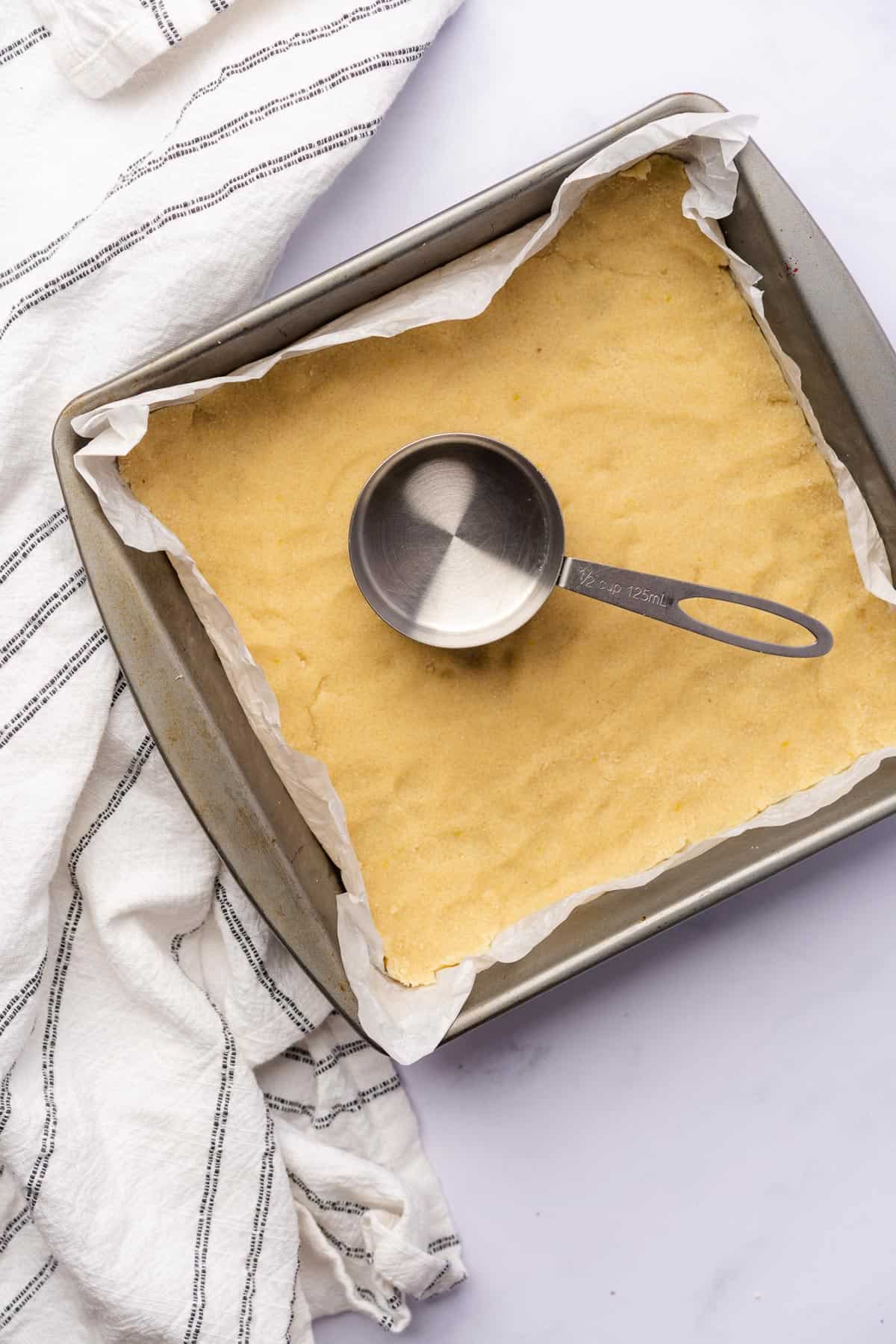 raw shortbread crust made from almond flour being pressed into a 9x9 inch pan with white parchment paper and a measuring cup flattening the dough