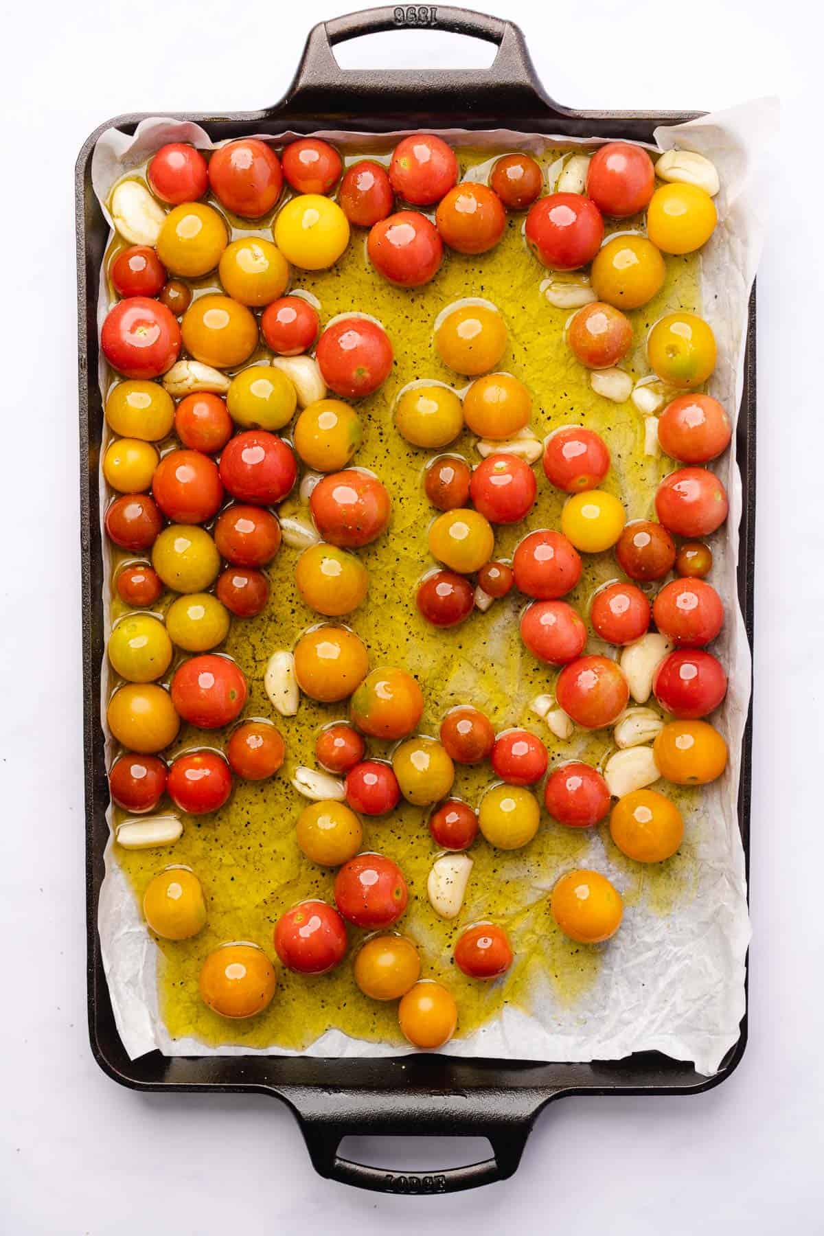 red, yellow and orange. cherry tomatoes with garlic cloves and olive oil on a parchment lined baking sheet