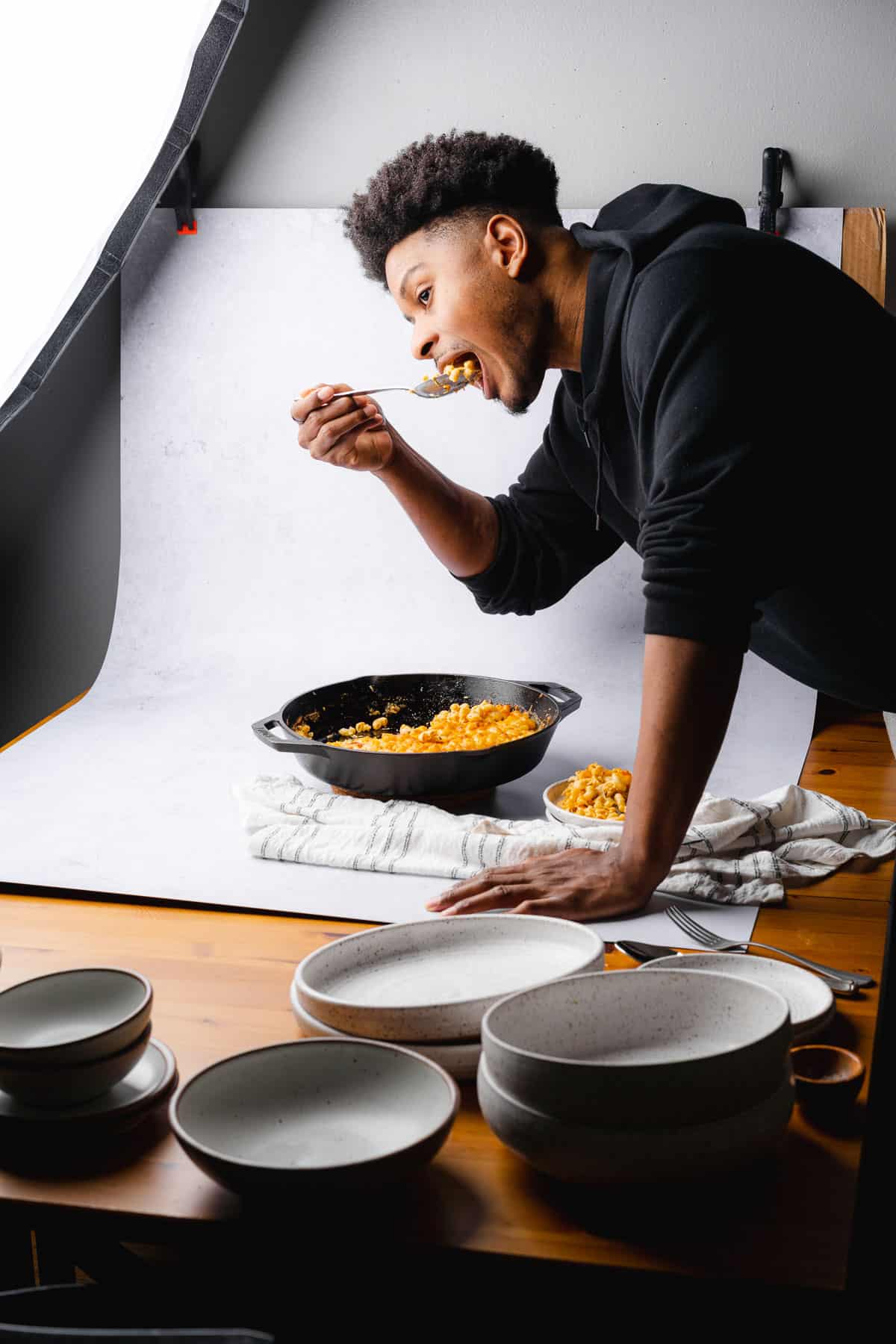 man leaning over a photography set to eat mac and cheese by the spoonful from a cast iron skillet