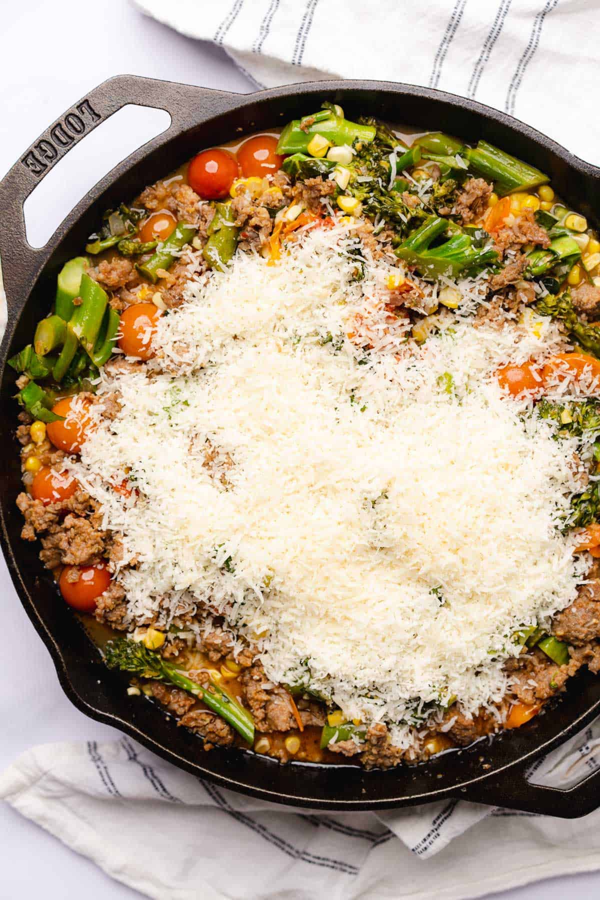 adding parmesan cheese to a skillet full of Italian sausage, corn, broccolini and tomatoes