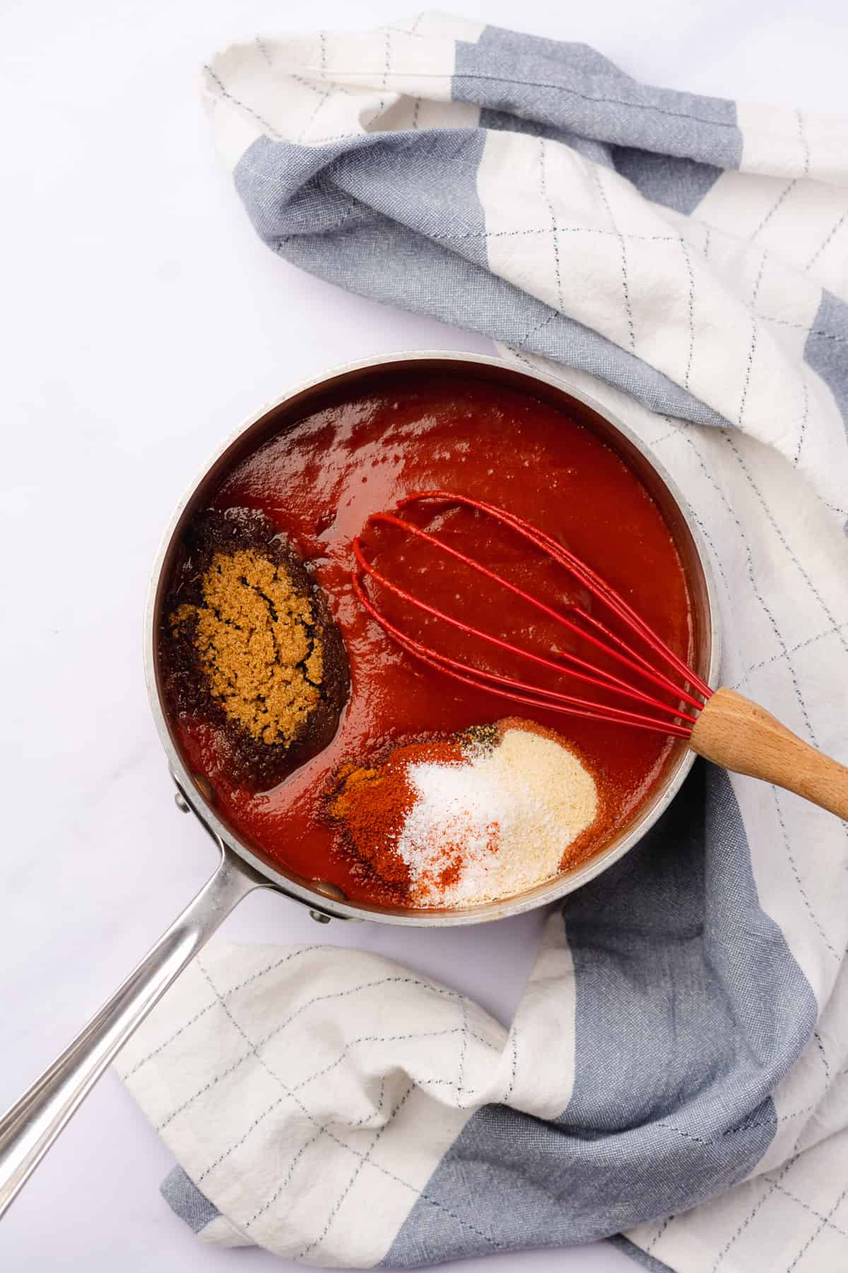 sweetener and seasonings added to tomato paste mixture in a saucepan with a red silicone whisk