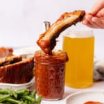 dipping delicious keto ribs into a jar of bbq sauce with beer and green beans on the scene