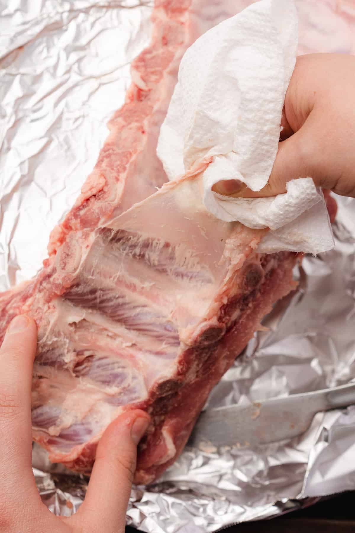 hand with paper toweling pulling back baby back ribs membrane